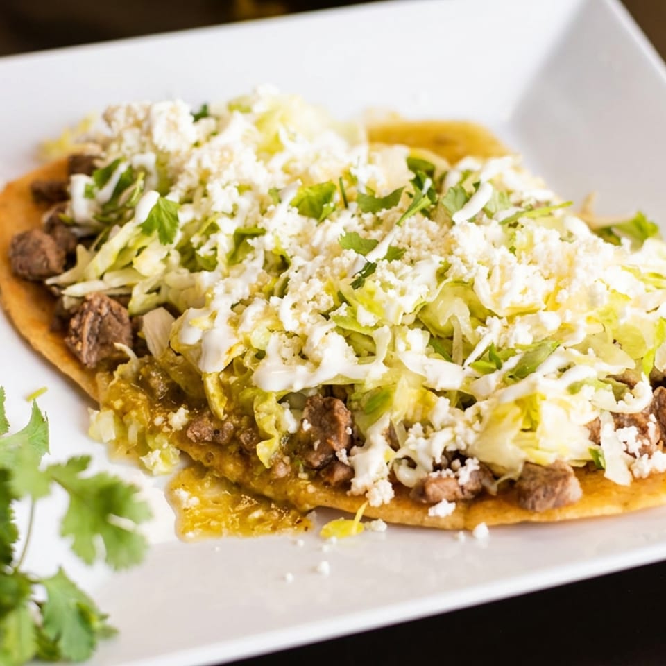 Savory tostada topped with seasoned meat, shredded cabbage, crumbled cheese, and sauces, with cilantro and peppers.