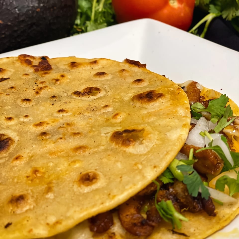 Two tacos with meat, onions, and cilantro on a white plate, with avocado and tomato in the background.
